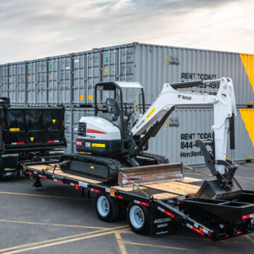 Herc Rentals truck with a Bobcat excavator loaded on a trailer in front of a stack of Herc Rentals containers, captured by Coldea Productions.