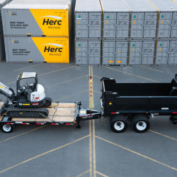 Herc Rentals truck with a Bobcat excavator loaded on a trailer in front of a stack of Herc Rentals containers, captured by Coldea Productions.