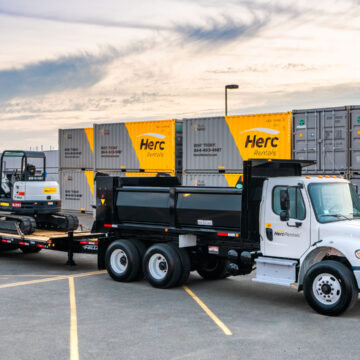 A Herc Rentals truck and trailer carrying a mini excavator, with storage containers in the background, showcasing Coldea Productions' commercial photography services.