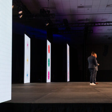 Convention-Photography Wide stage view at a conference with LED screens displaying visual designs and two speakers on stage.