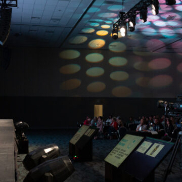 Convention-Photography Camera operator capturing two speakers on stage at a large conference event, with an attentive audience and vibrant lighting.