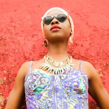 Stylish woman in sunglasses standing against a textured red wall, wearing vibrant clothing and statement jewelry, photographed by Coldea Productions.