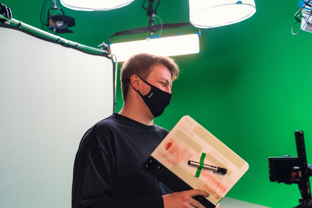 A masked video production crew member holding a clapperboard on a green screen set.