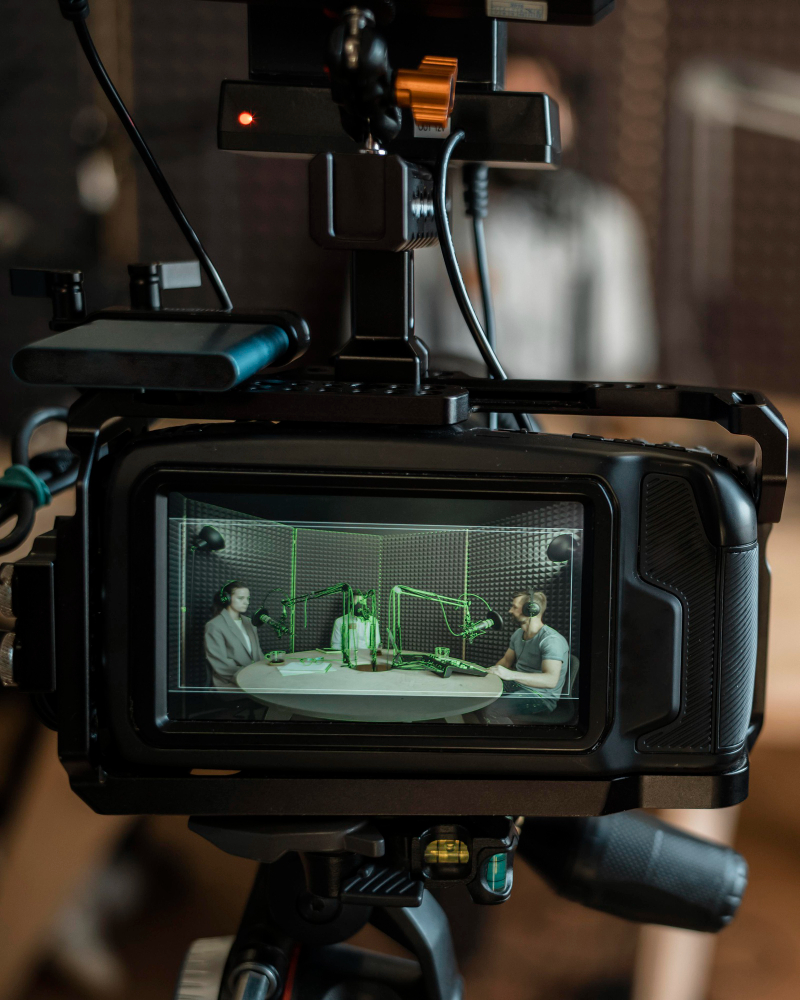 Camera capturing a podcast recording in a studio with three people around a table using professional microphones.