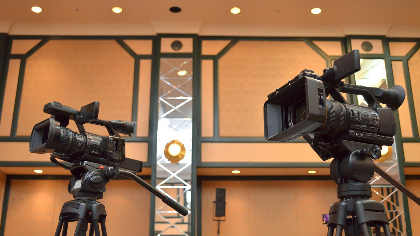Two professional video cameras on tripods, set up in an elegant conference venue, ready to film an event.