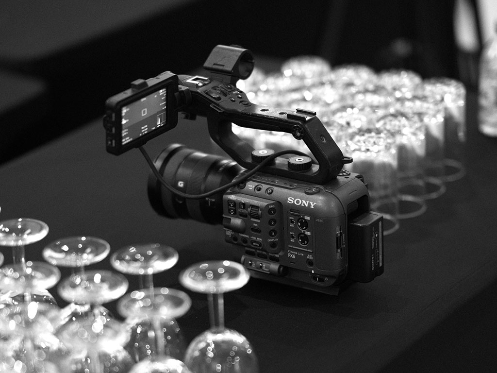 A Sony FX6 video camera setup on a table with rows of wine glasses in the background, showcasing professional equipment used in convention and conference videography.