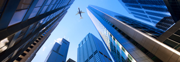 View of skyscrapers from the ground with an airplane flying overhead, representing corporate growth and expansion.