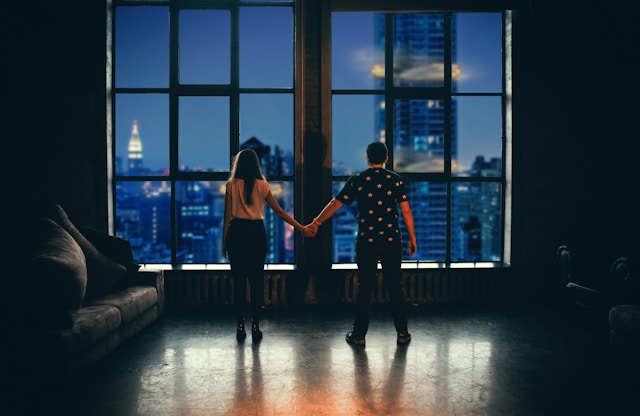 A couple holding hands while gazing at a city skyline through large loft windows, showcasing urban living and modern luxury.