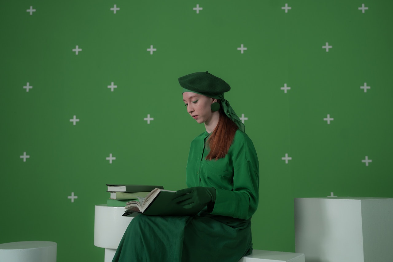 A woman dressed entirely in green, including a beret and gloves, sits reading a book against a green screen backdrop with tracking markers, ready for visual effects compositing.