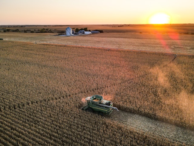 Aerial view of a combine harvester working in a large cornfield at sunset, with a farmhouse and silos in the background.