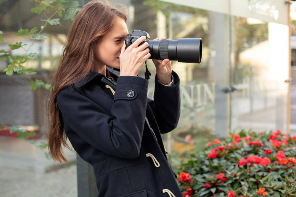 A young female photographer taking a photo outdoors, holding a professional camera with a large zoom lens.