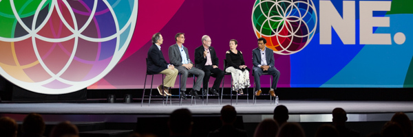 A group of five people seated on stage in front of a colorful, geometric backdrop featuring the word "ONE".