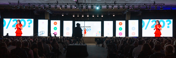 A large-scale corporate training event with a presenter on stage, highlighted by vibrant screens displaying engaging visuals, with a professional camera capturing the moment.