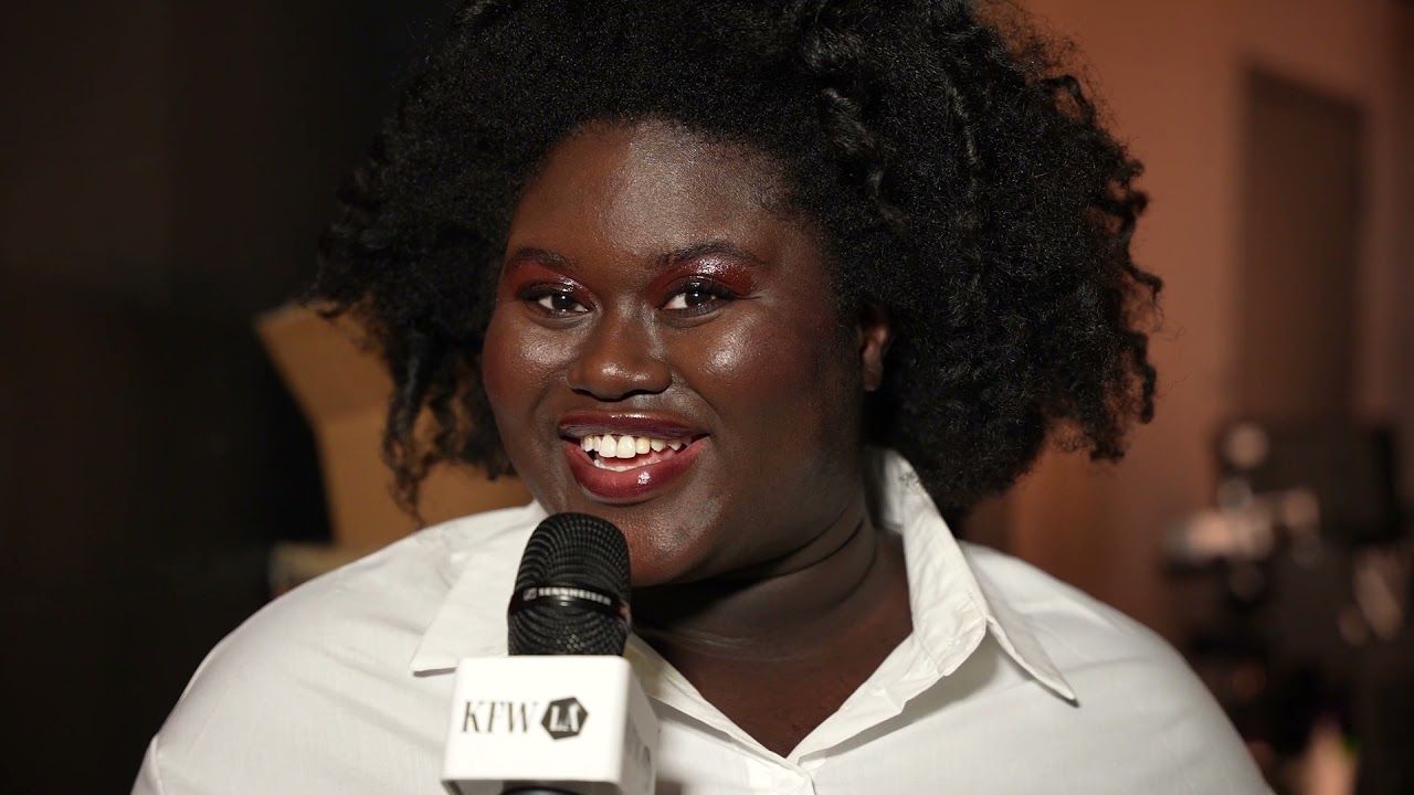A joyful speaker with natural hair and bright makeup recaps the events of the day at Kornit Fashion Week LA 2021. She is holding a microphone with a KFW LA logo, expressing enthusiasm for the day's highlights.