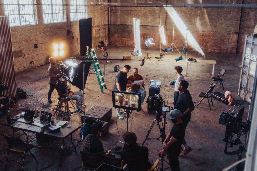 Coldea Productions crew preparing for a film shoot in a spacious industrial studio. The team is setting up multiple cameras, lights, and other production equipment. Several crew members are working with the talent seated in the center, while others adjust the lighting and review footage on monitors and laptops.