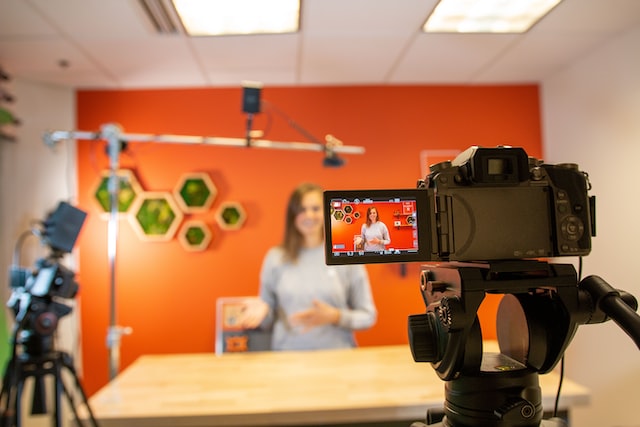 A professional camera captures a woman standing behind a desk in a brightly lit studio with orange walls. The screen of the camera shows the woman presenting, while another camera and microphone are set up around her.