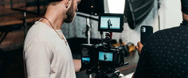 Videographers capturing live footage of a subject in a professional studio, with the camera feed visible on a monitor and a smartphone for live streaming.