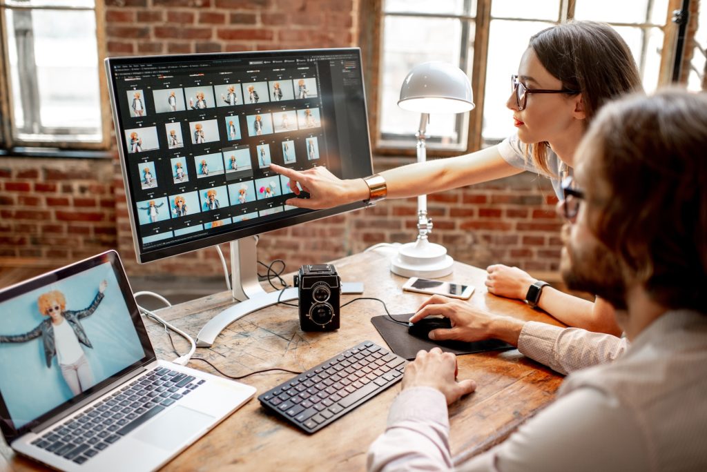 Two people reviewing and editing professional photos on a large desktop monitor in a creative workspace, with photography equipment on the desk.