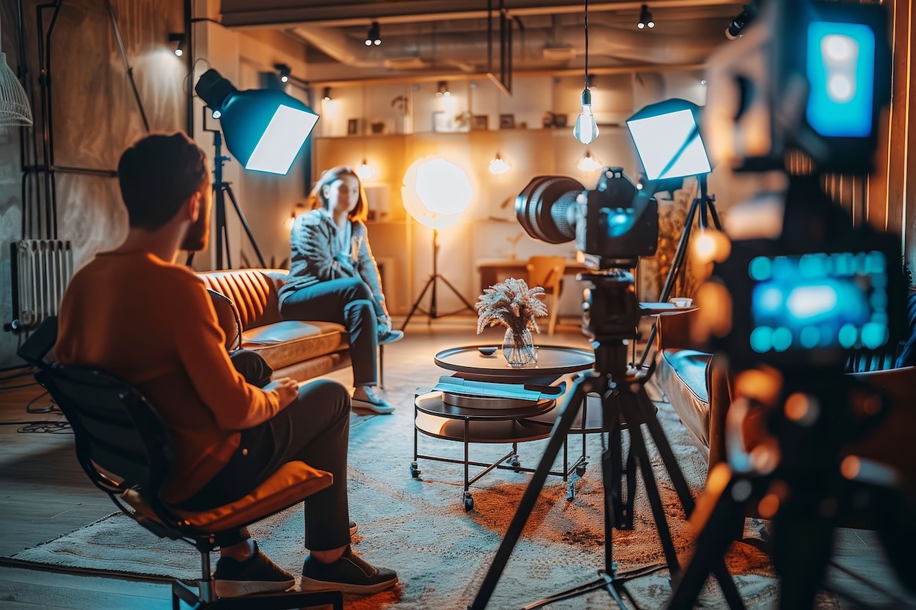 Behind-the-scenes of a professional interview setup, with lighting, cameras, and a crew member capturing a seated interview subject in a cozy studio.