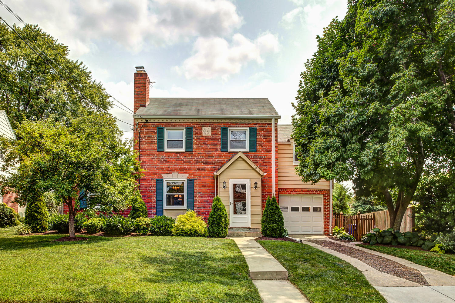 A beautifully landscaped two-story brick home with a paved driveway, vibrant greenery, and a clear sky, captured to represent real estate photography.