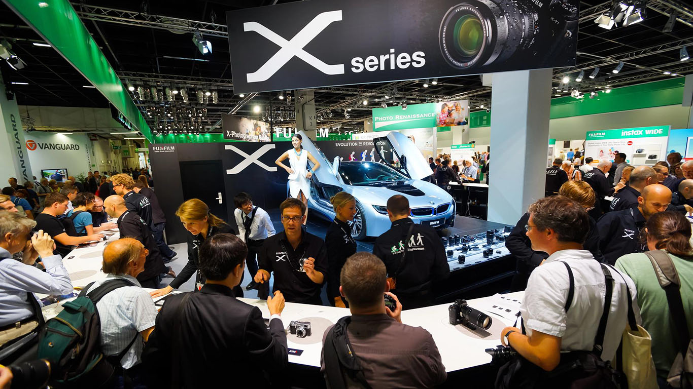 A bustling trade show booth featuring the Fujifilm X Series, with attendees engaging with product displays and staff, showcasing dynamic trade show activities.