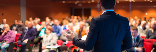 A professional speaker addressing a large audience during a corporate training event in a modern conference hall.