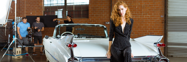 A woman with curly red hair wearing a black suit jacket is standing in front of a classic white car in what appears to be a garage or vehicle showroom.