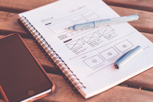Notebook with website wireframes and smartphone on a wooden desk, symbolizing visual consistency and design planning for building trust online.